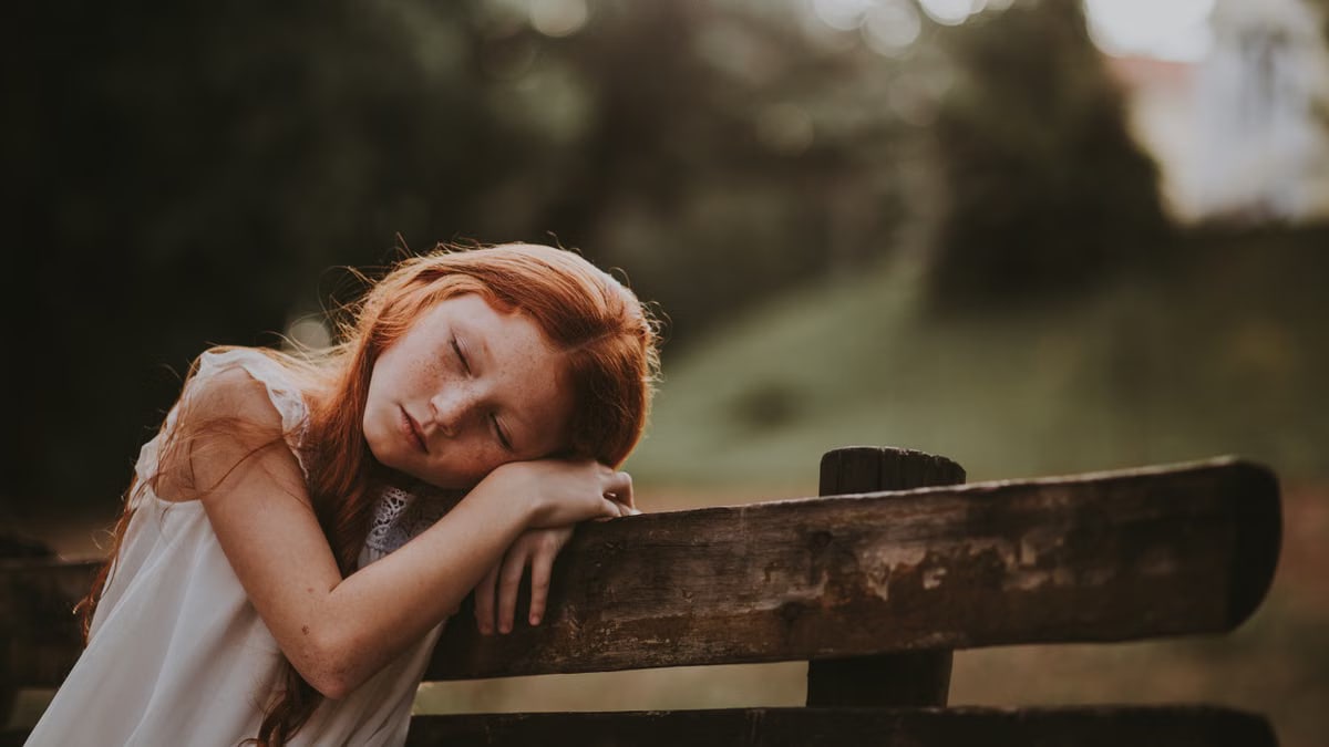 A young girl sleeps peacefully on a wooden bench outdoors, surrounded by nature.